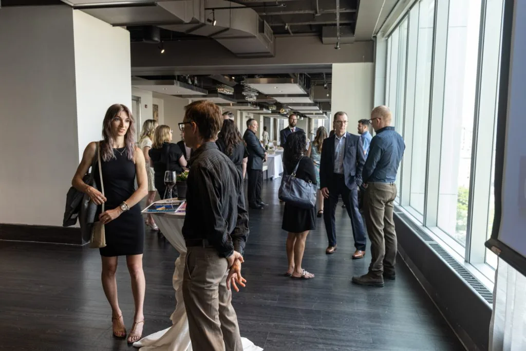 Attendees mingling at an evening reception in a glass-walled venue, some holding wine glasses